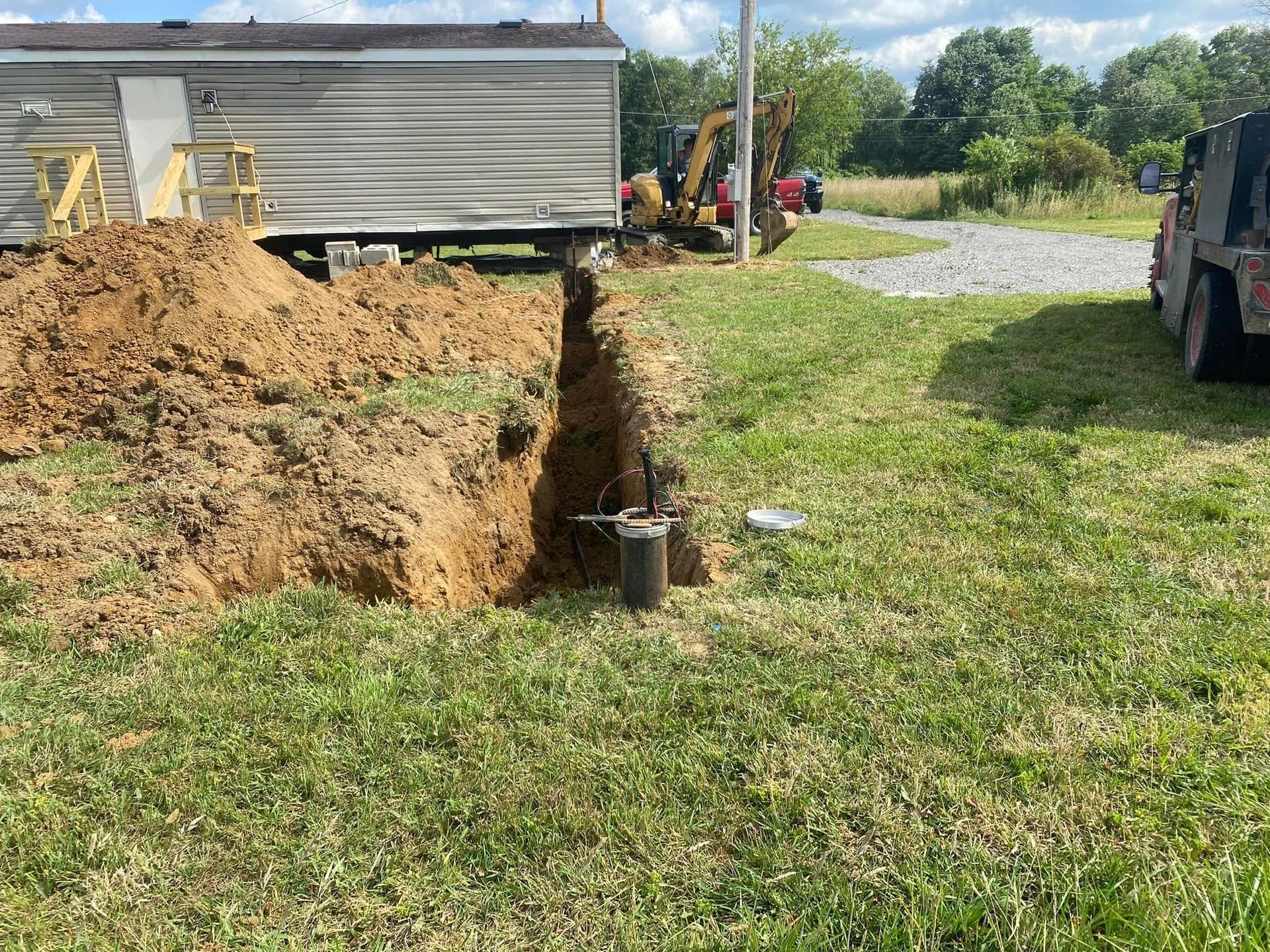A trench dug in a grassy yard near a mobile home, possibly for utility lines, with an excavator in the background.