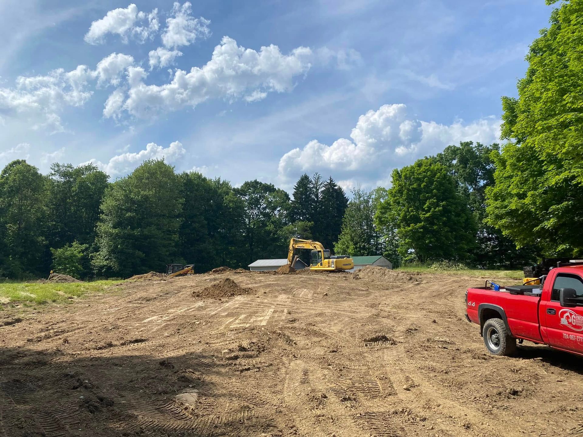 A cleared construction site with a yellow excavator, trees, and a red pickup truck under a blue sky with fluffy clouds.