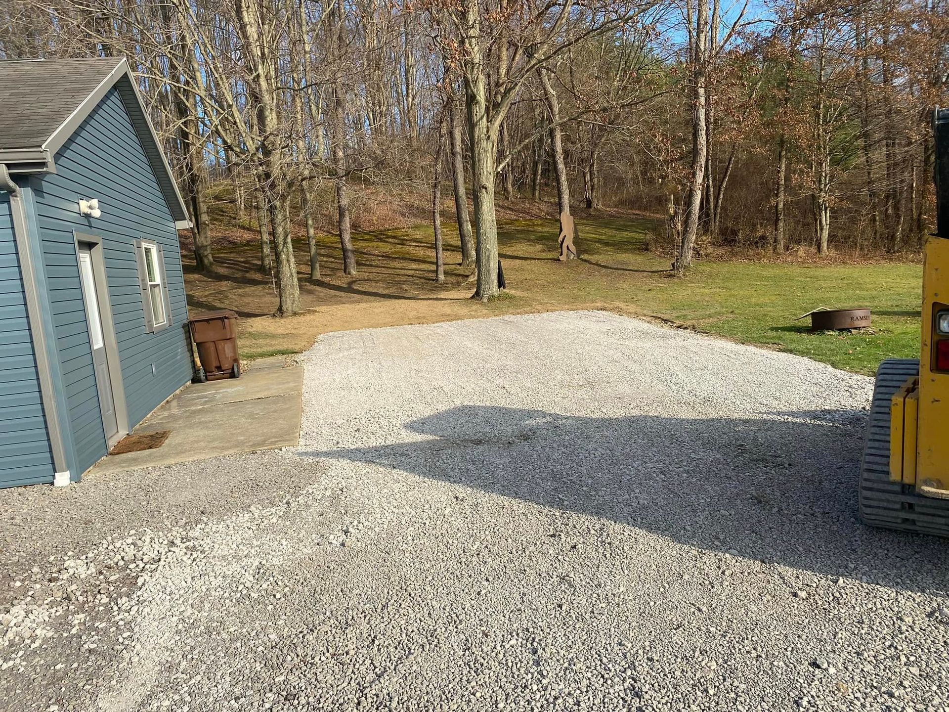 A gravel driveway next to a blue house, with a small tractor on the right. Trees and a grassy hill are in the background.