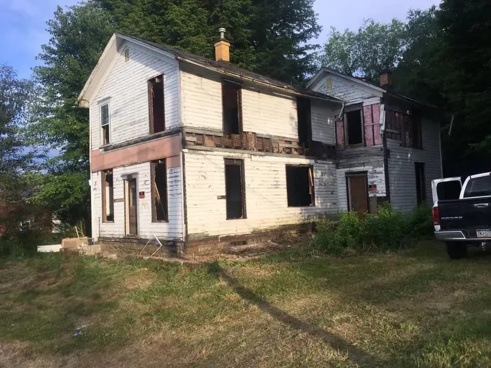Dilapidated two-story house with missing windows and siding, set on a grassy hill. A truck is parked nearby.