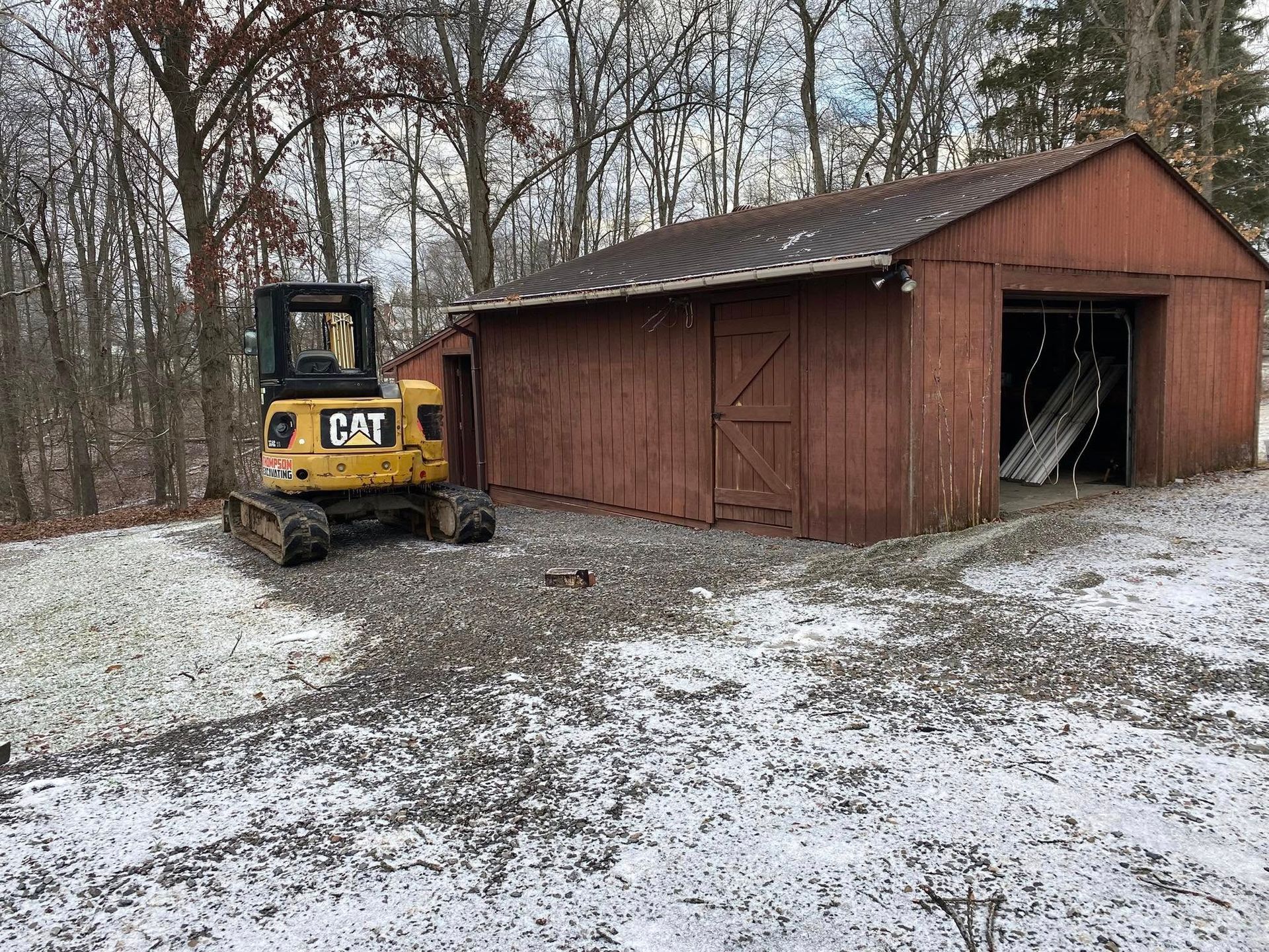 Yellow CAT excavator parked near a weathered red barn in a snowy, wooded area. The barn door is open.