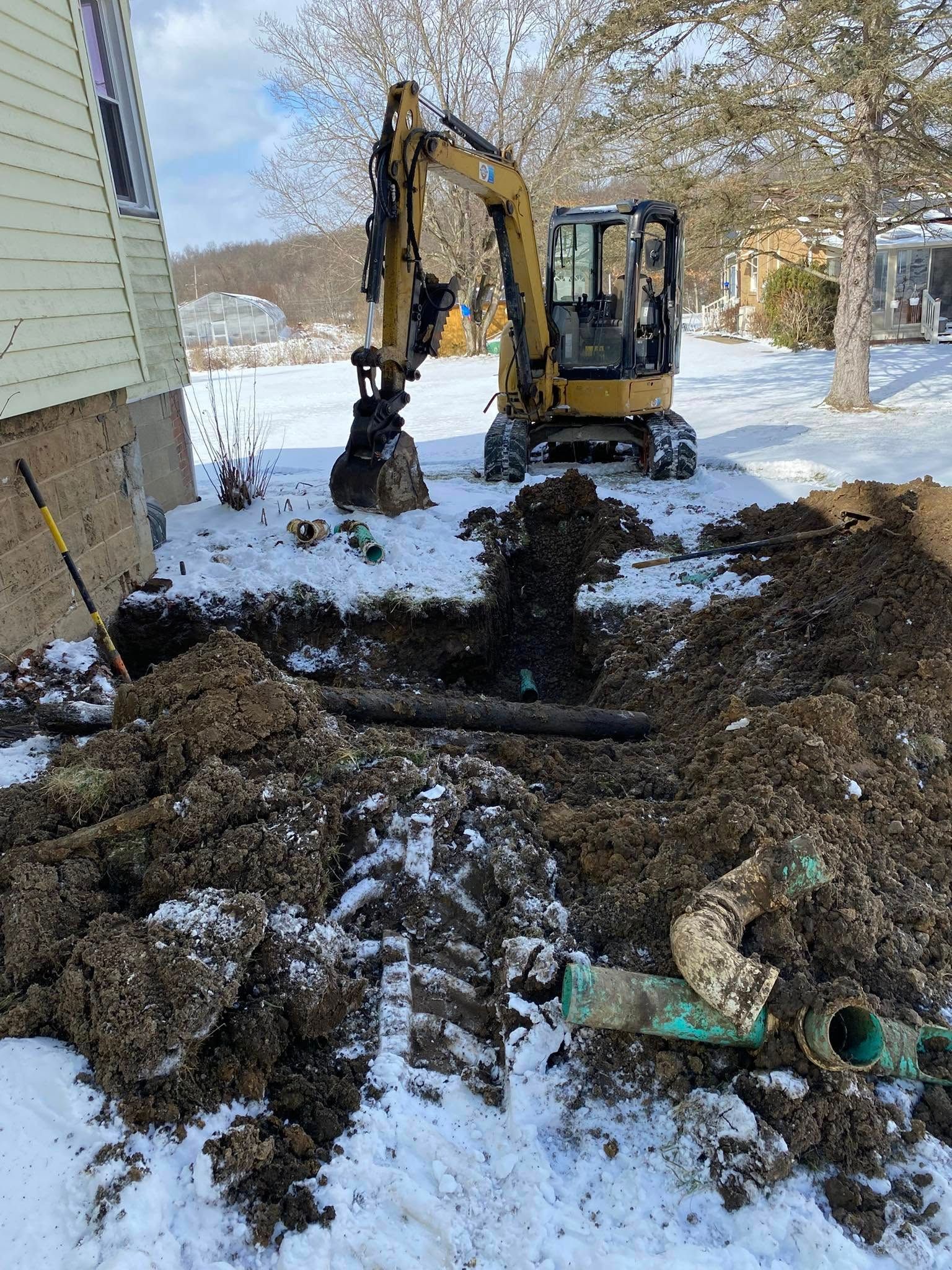 A small excavator digging in a yard covered in snow, near a house. Mud and pipes are visible, indicating plumbing work.
