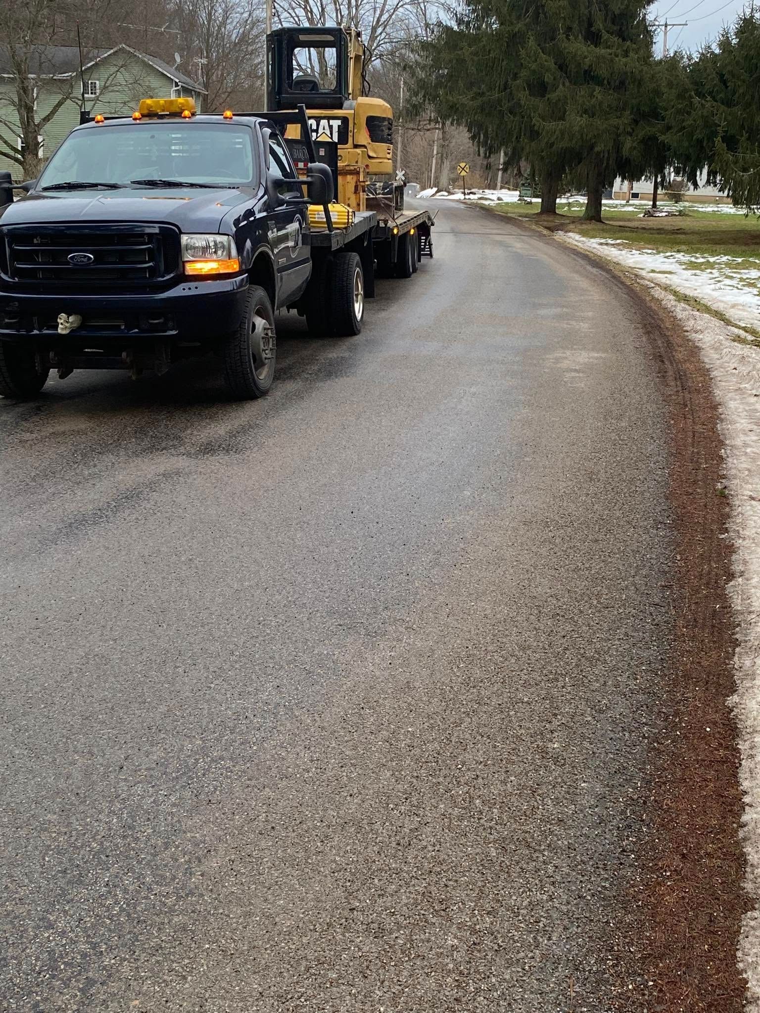 A dark blue truck with a Caterpillar machine on a flatbed trailer on a wet road. The road has ice on the shoulder.