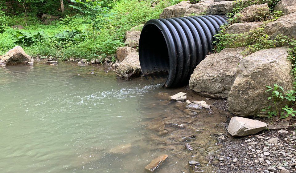 Black corrugated pipe emptying water into a stream, rocks and green foliage surrounding.