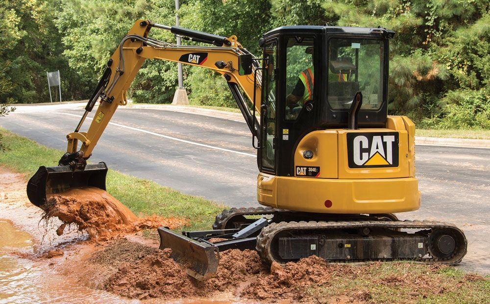 A yellow Caterpillar mini excavator digging in muddy soil next to a road.