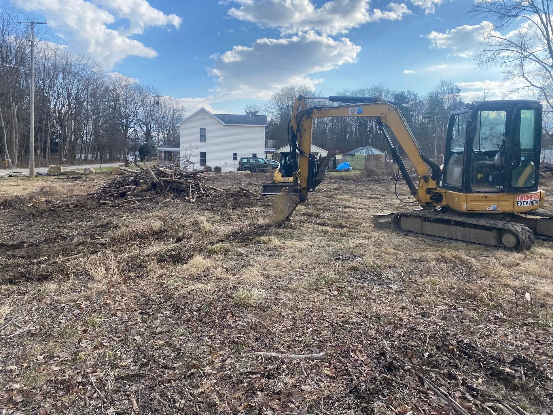 A yellow excavator clearing brush on a brown field with a white house in the background.