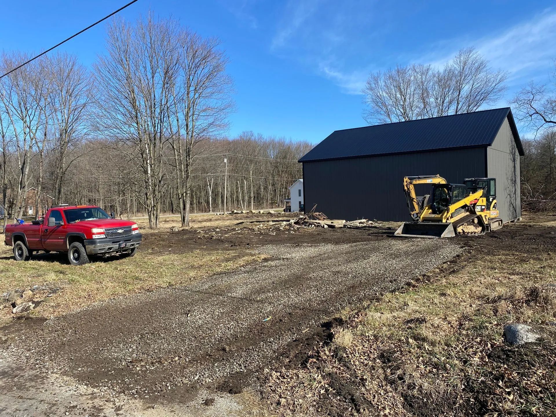A red pickup truck and yellow skid steer are parked near a dark gray barn on a dirt lot with a gravel pathway.