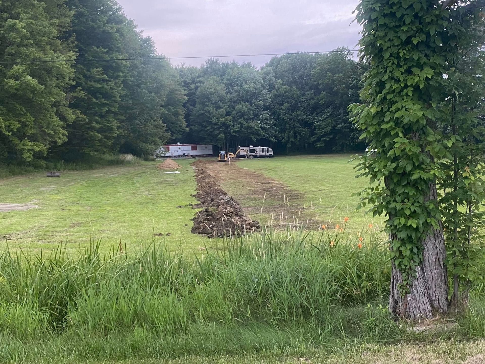 A field with a trench being dug. In the background, a mobile home is visible. A tree with vines is in the foreground.