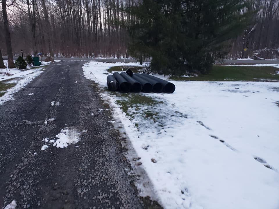 Black drainage pipes lie on snow-covered grass beside a dark gravel driveway.