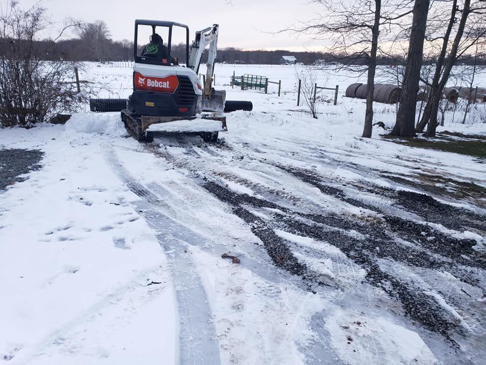 A Bobcat excavator plows snow from a driveway in a rural setting; the operator is visible.