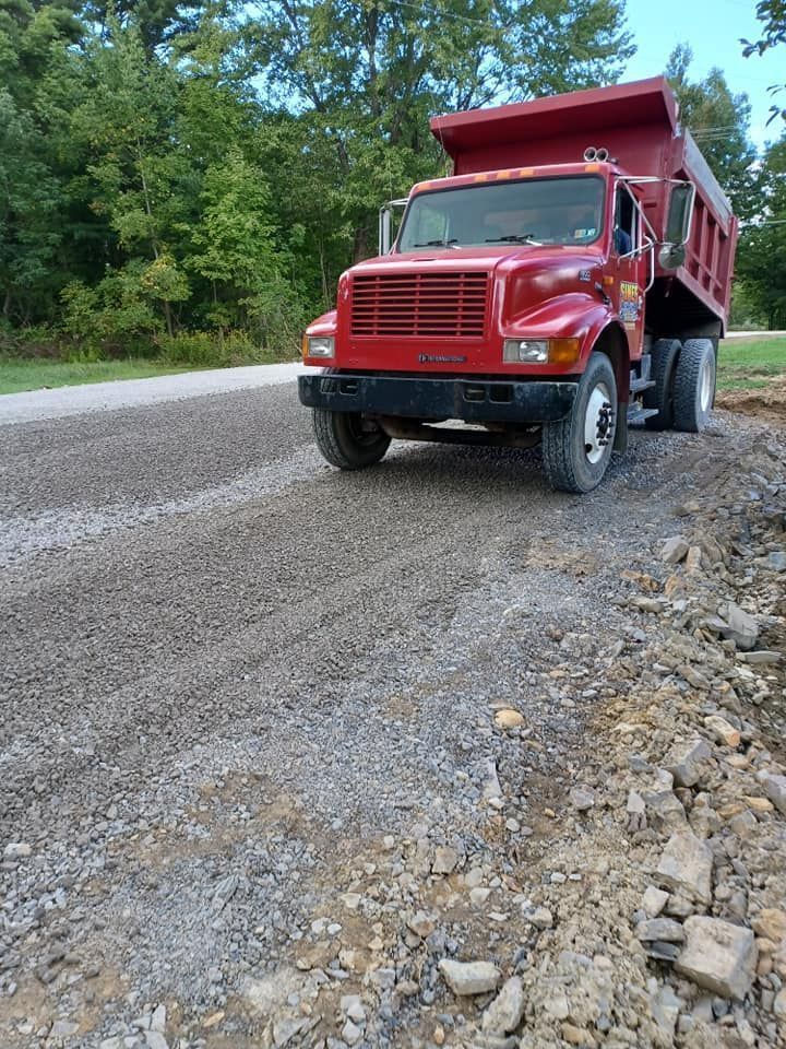 Red dump truck on a gravel road, likely spreading material for road construction. Trees and grass in the background.
