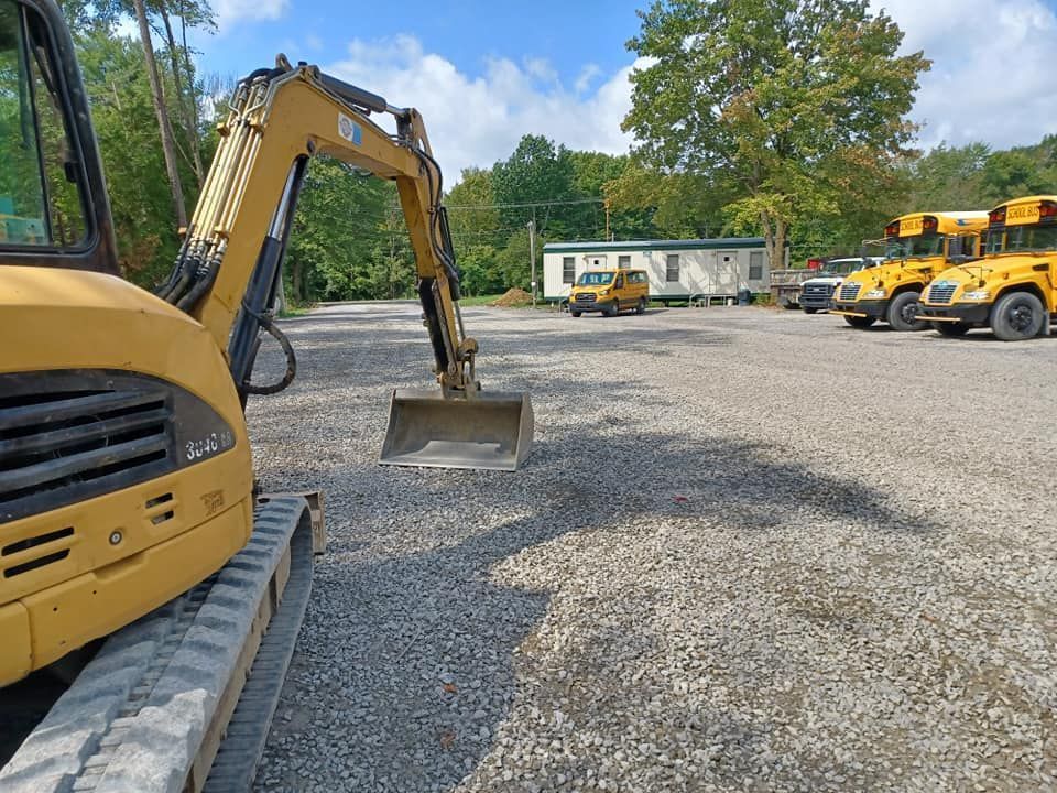 Yellow excavator in a gravel lot, with school buses and a mobile office in the background on a sunny day.