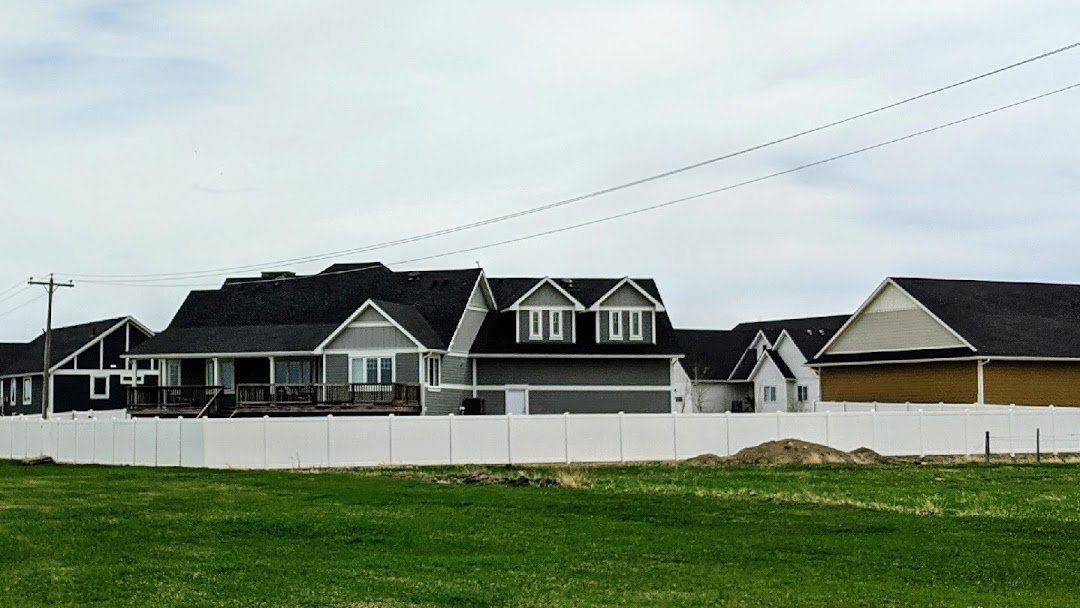 Suburban homes with dark roofs, white fence, green grass, and overcast sky.