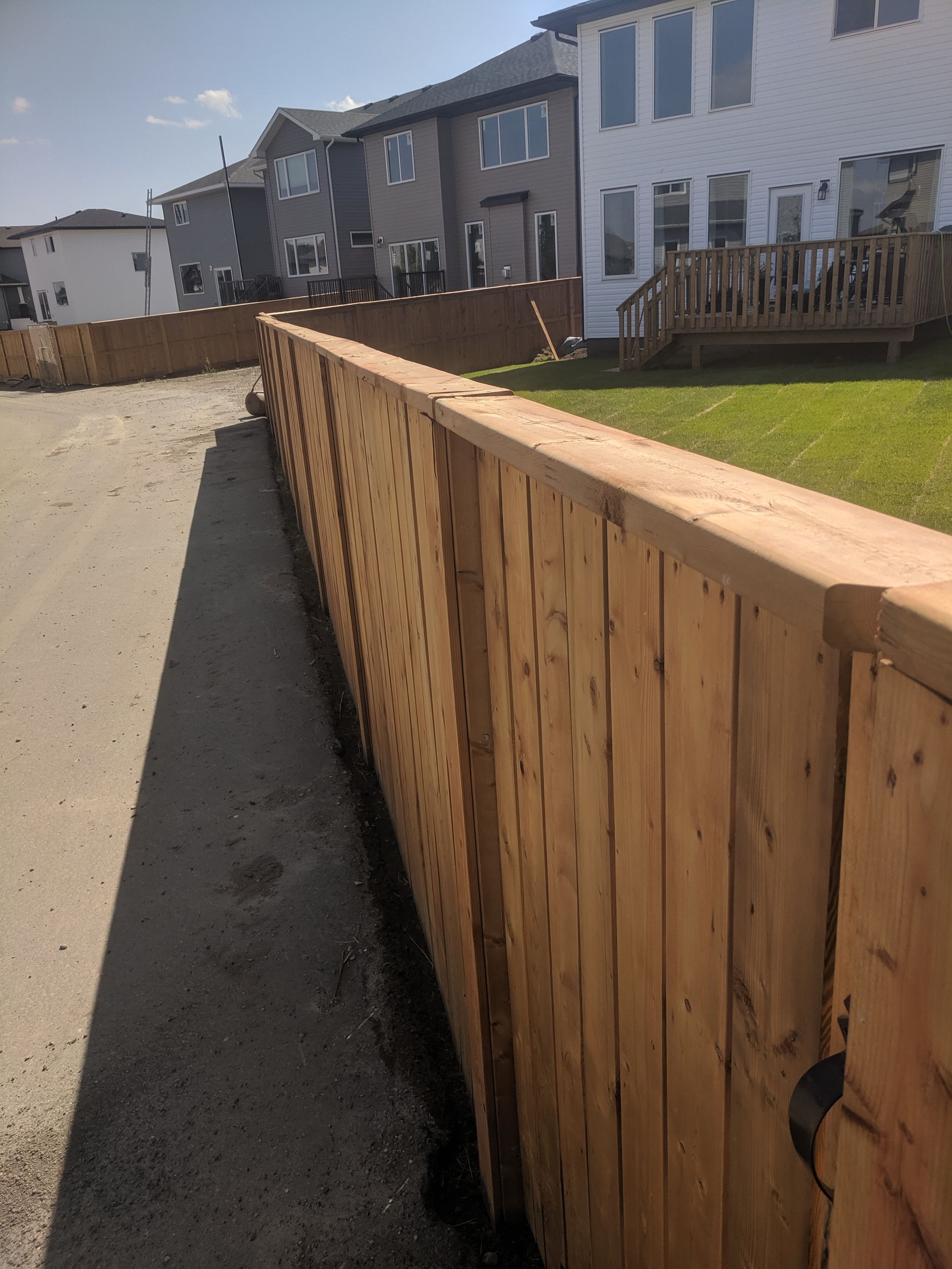 Wooden fence along a gravel driveway in a suburban neighborhood, houses in the background.