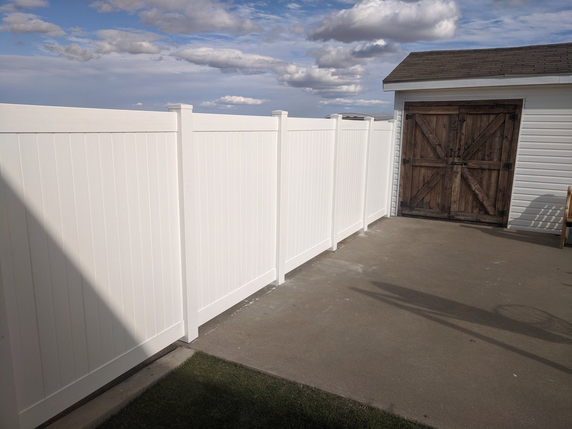 White vinyl fence bordering a concrete patio, with a brown-doored shed in the background under a cloudy sky.