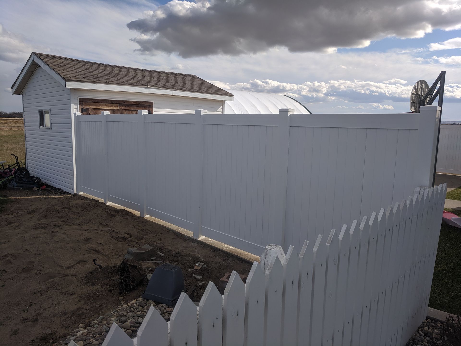 White picket fence transitioning to tall white privacy fence near a shed, under a cloudy sky.
