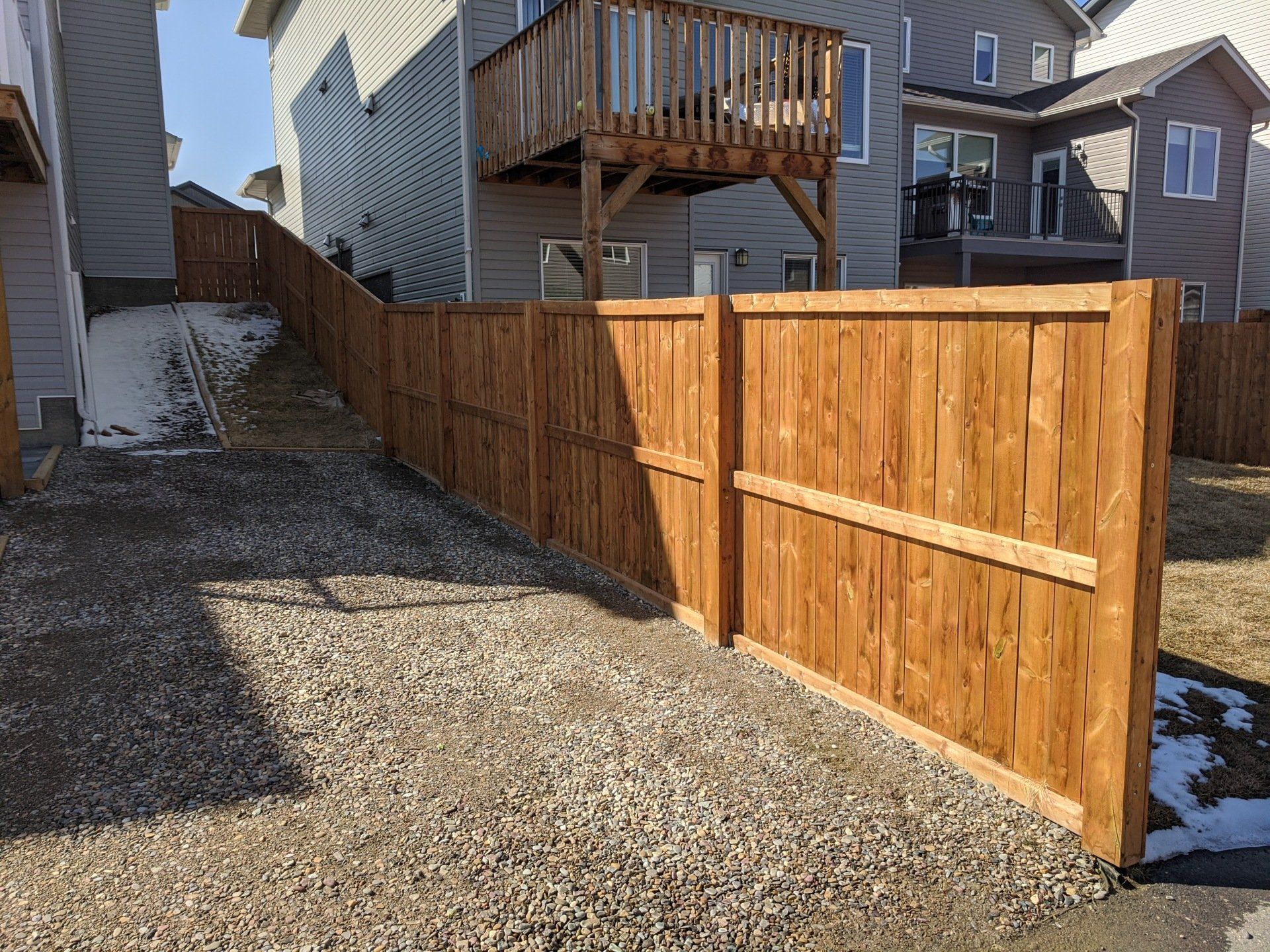Wooden fence on gravel between houses, a deck visible on the house in the background.