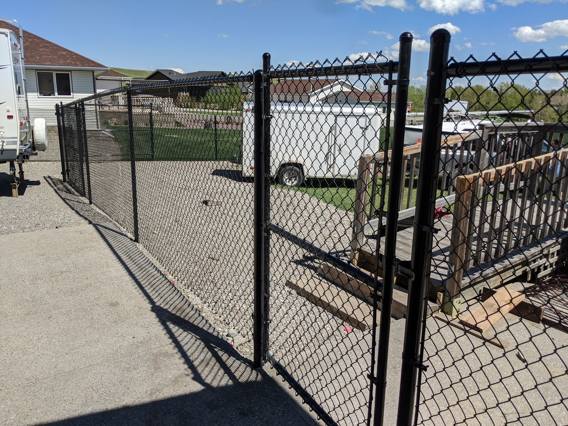 Black chain-link fence encloses a yard with a trailer, deck, and house on a sunny day.