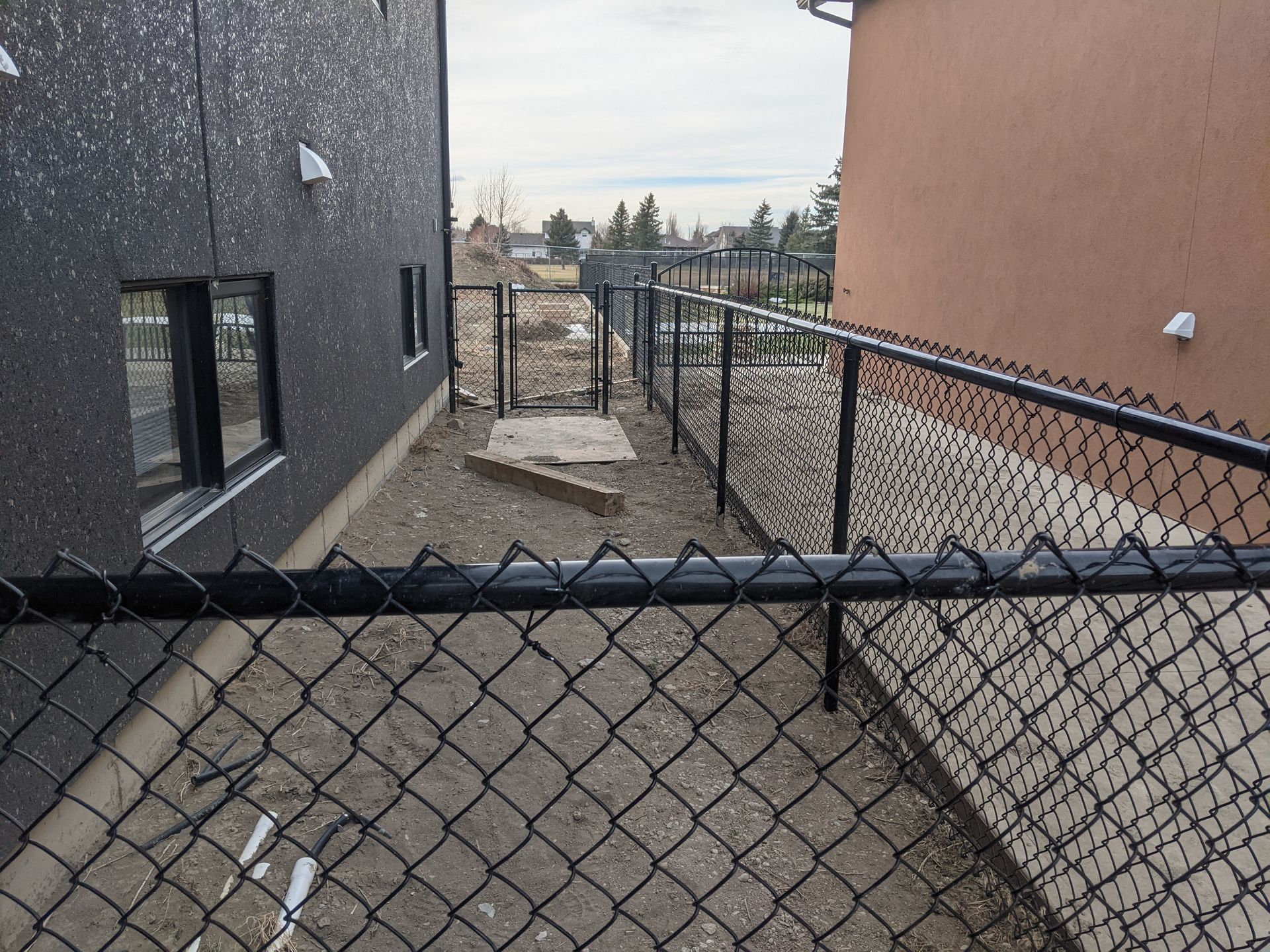 Black chain-link fence between two buildings, leading to a gate. Dry dirt ground.