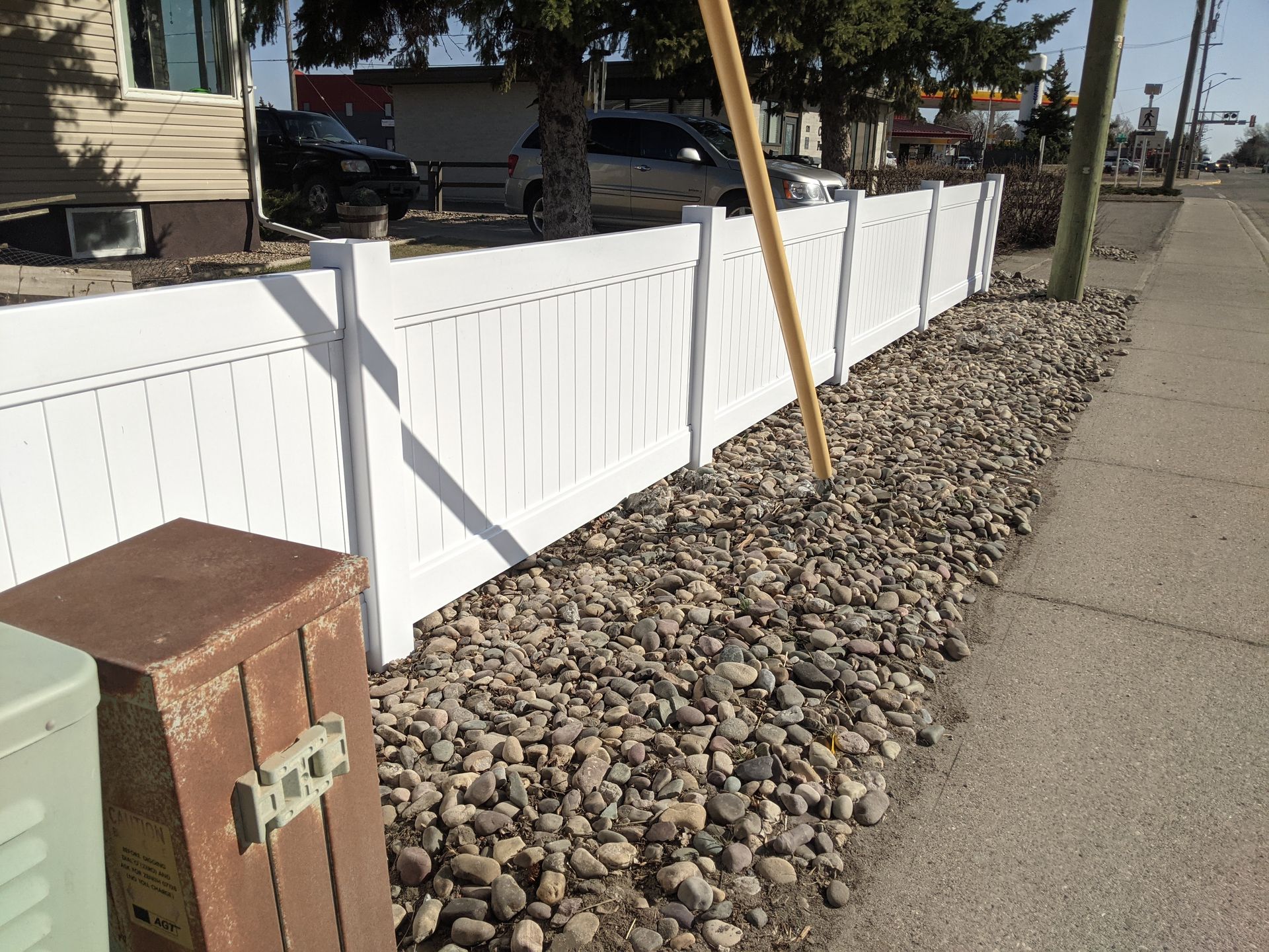 White picket fence along a sidewalk, with rocks in between, near a building and parked cars.