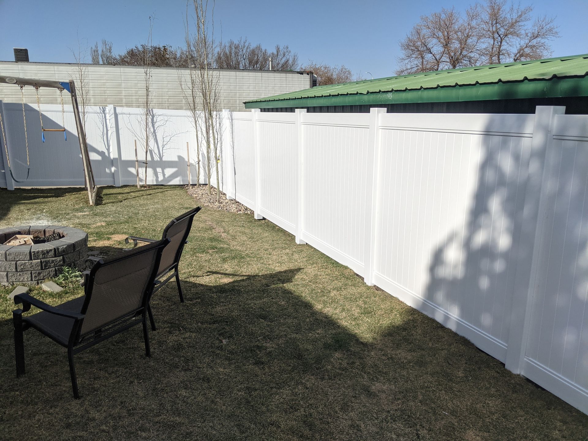White vinyl fence encloses a grassy yard with two chairs, a fire pit, and small trees under a blue sky.