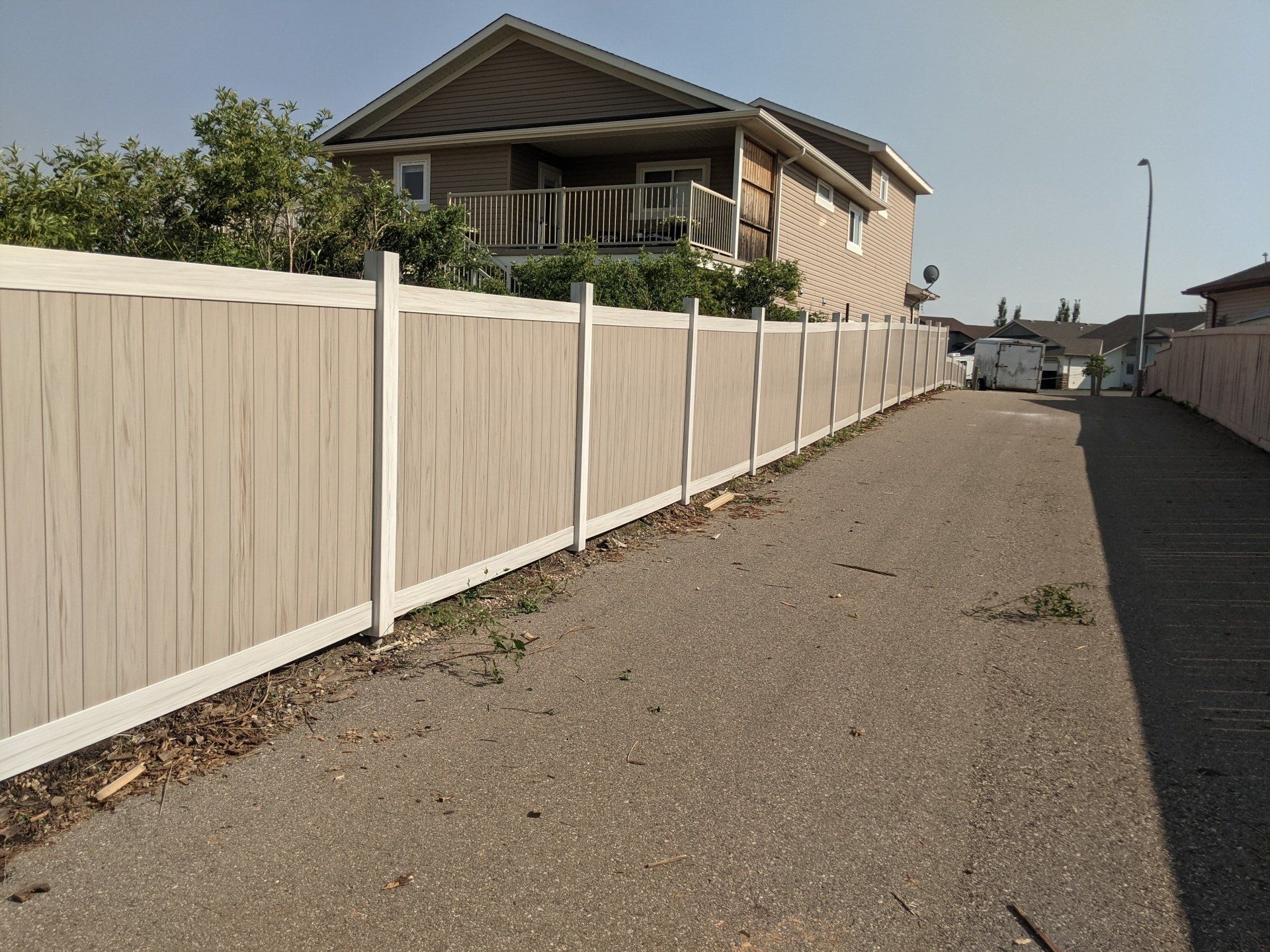 A gravel alley with a tan and white fence alongside a two-story house on a sunny day.