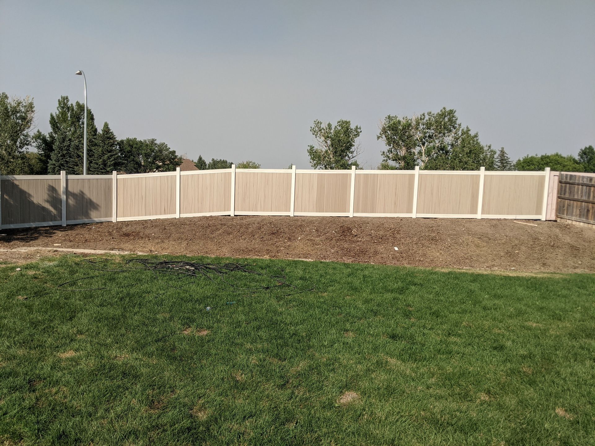 Tan and white vinyl fence along a grassy lawn, with trees and cloudy sky in the background.
