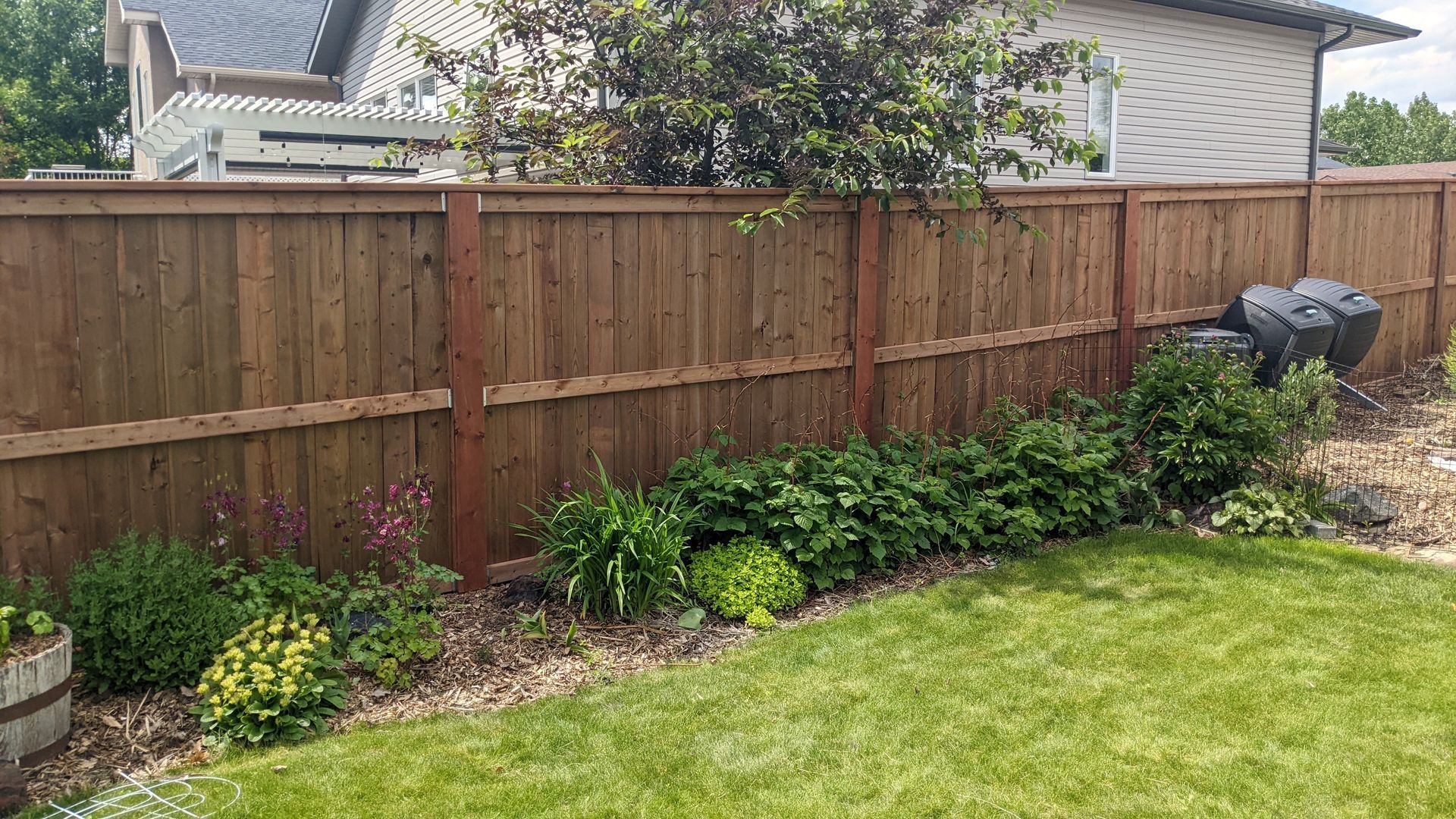 Wooden fence with a row of green bushes, flowers, and grass in front of a house.