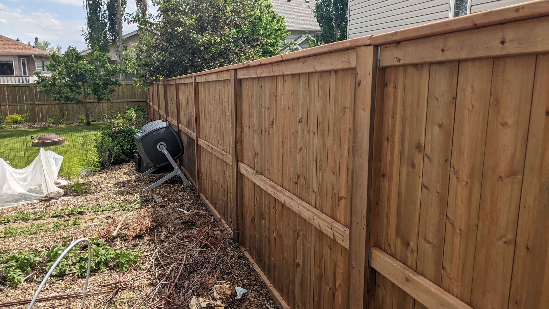 Wooden fence in a backyard, with brown boards and supporting beams, near a compost bin.