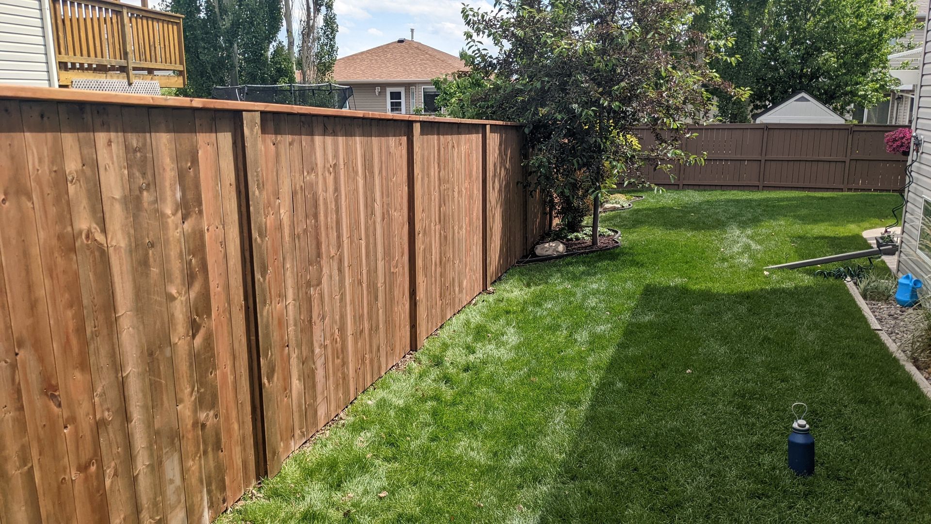 Wooden fence surrounds green grass in a backyard with a small tree and sprinklers.
