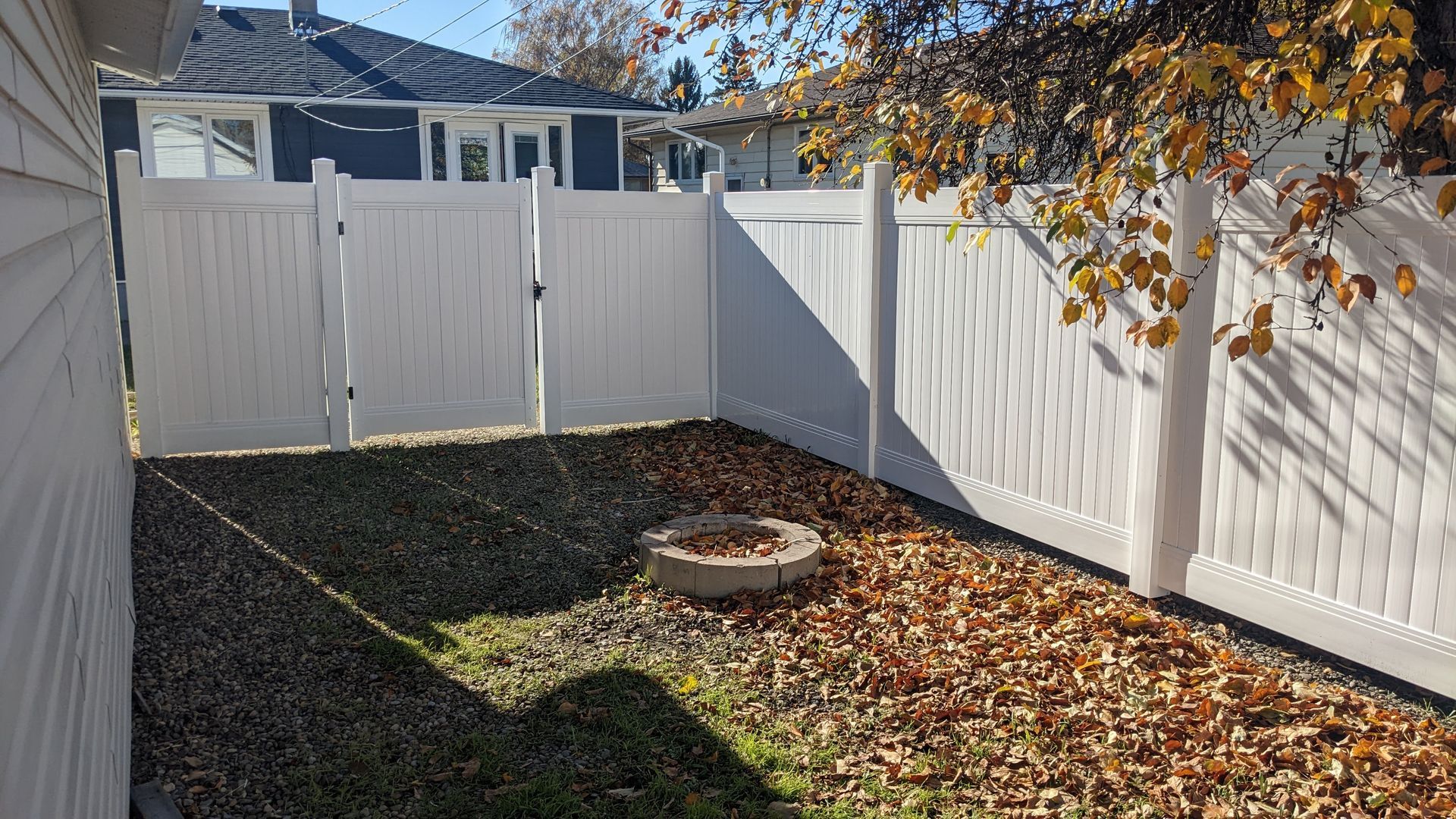 White fenced backyard with fire pit and fall leaves.