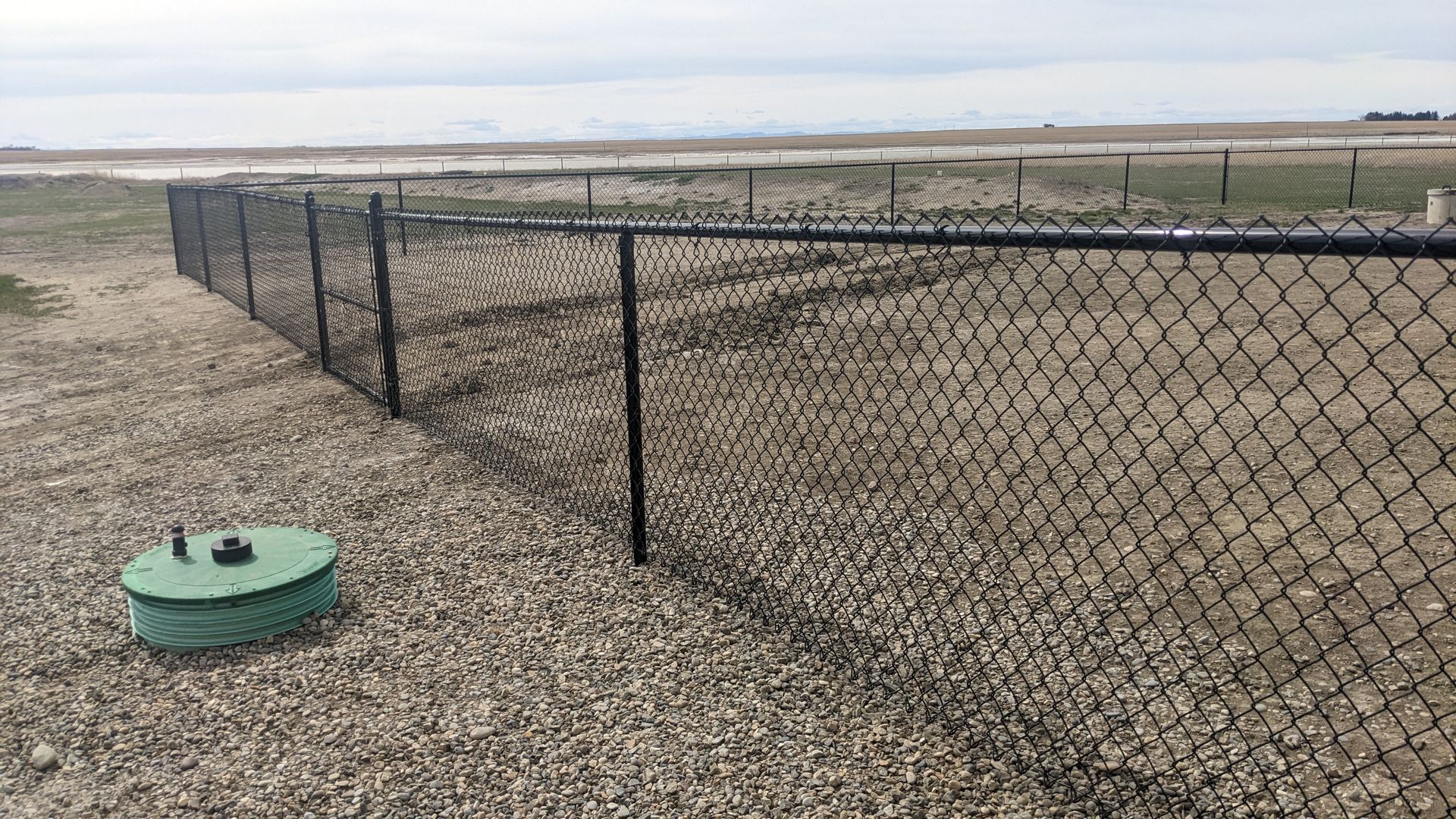 Black chain-link fence surrounds a gravel-covered area with a green utility cover in the foreground, outdoors.