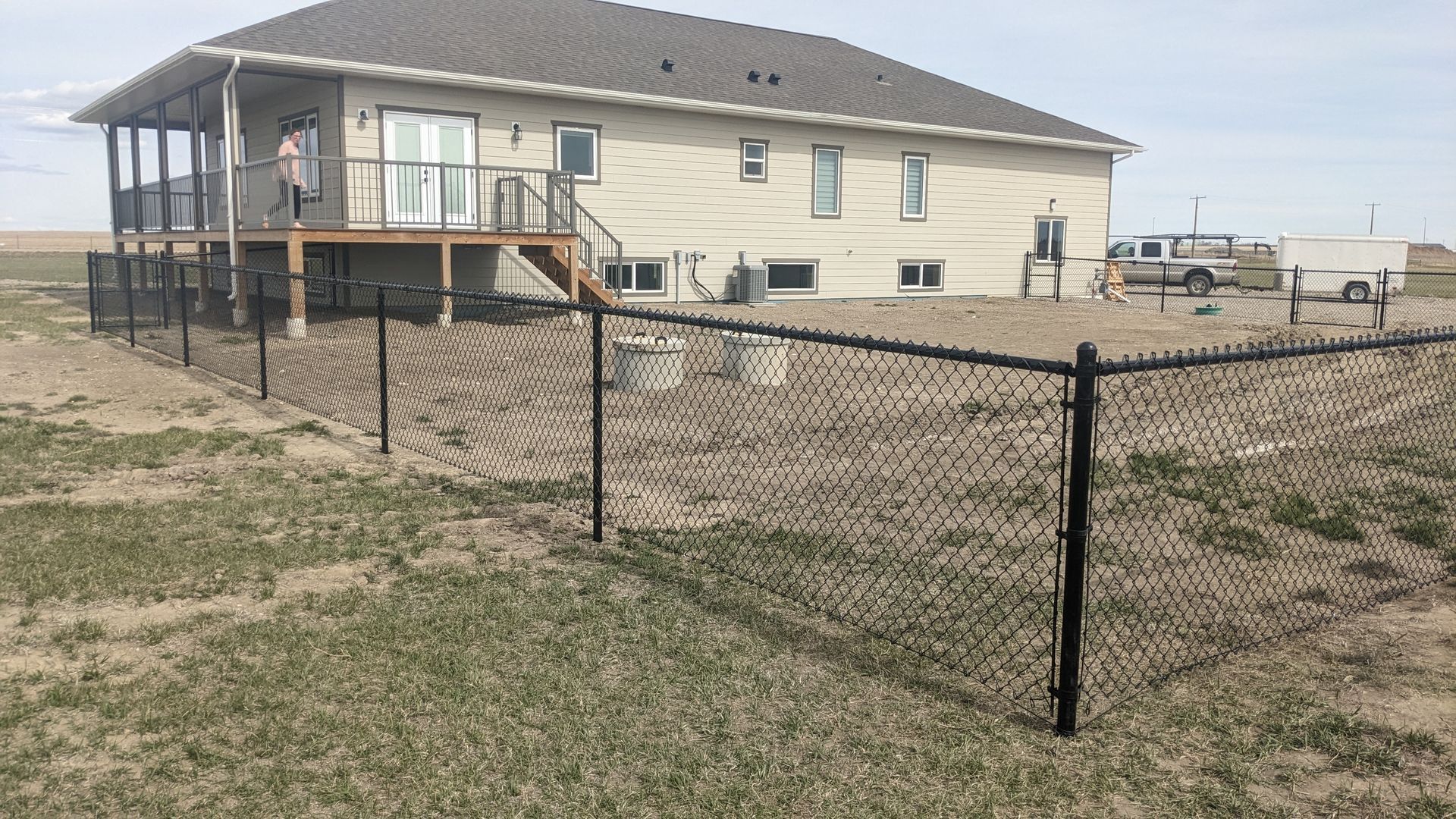 Black chain-link fence around a beige house with a deck, set in a grassy field.