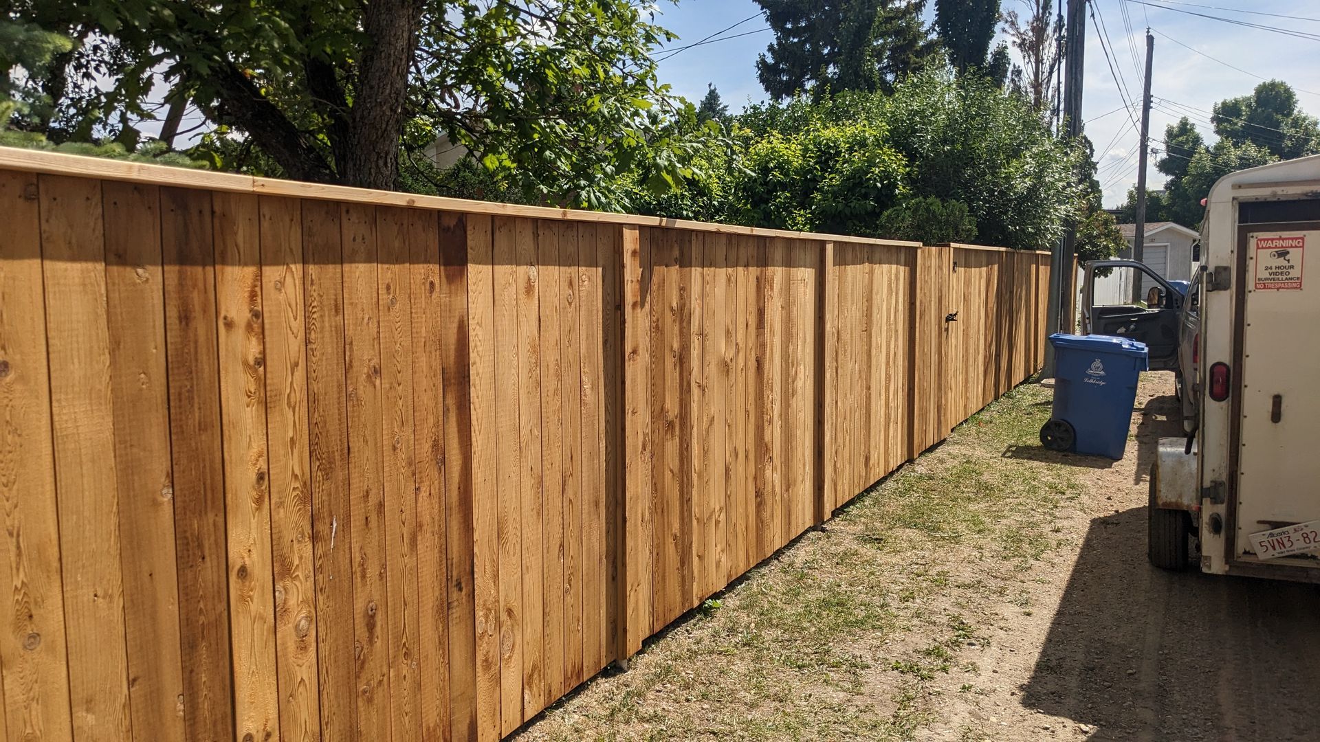 Wooden fence along a narrow gravel area, with a blue trash can and trailer visible.