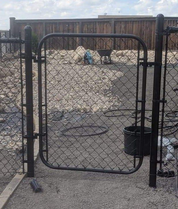 Black chain-link gate in a gravel yard with a wooden fence background.