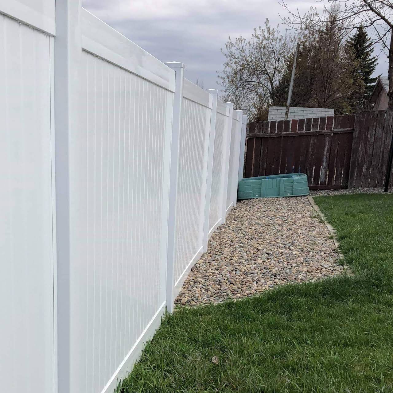 White vinyl fence bordering a grassy yard with a rock bed, under an overcast sky.