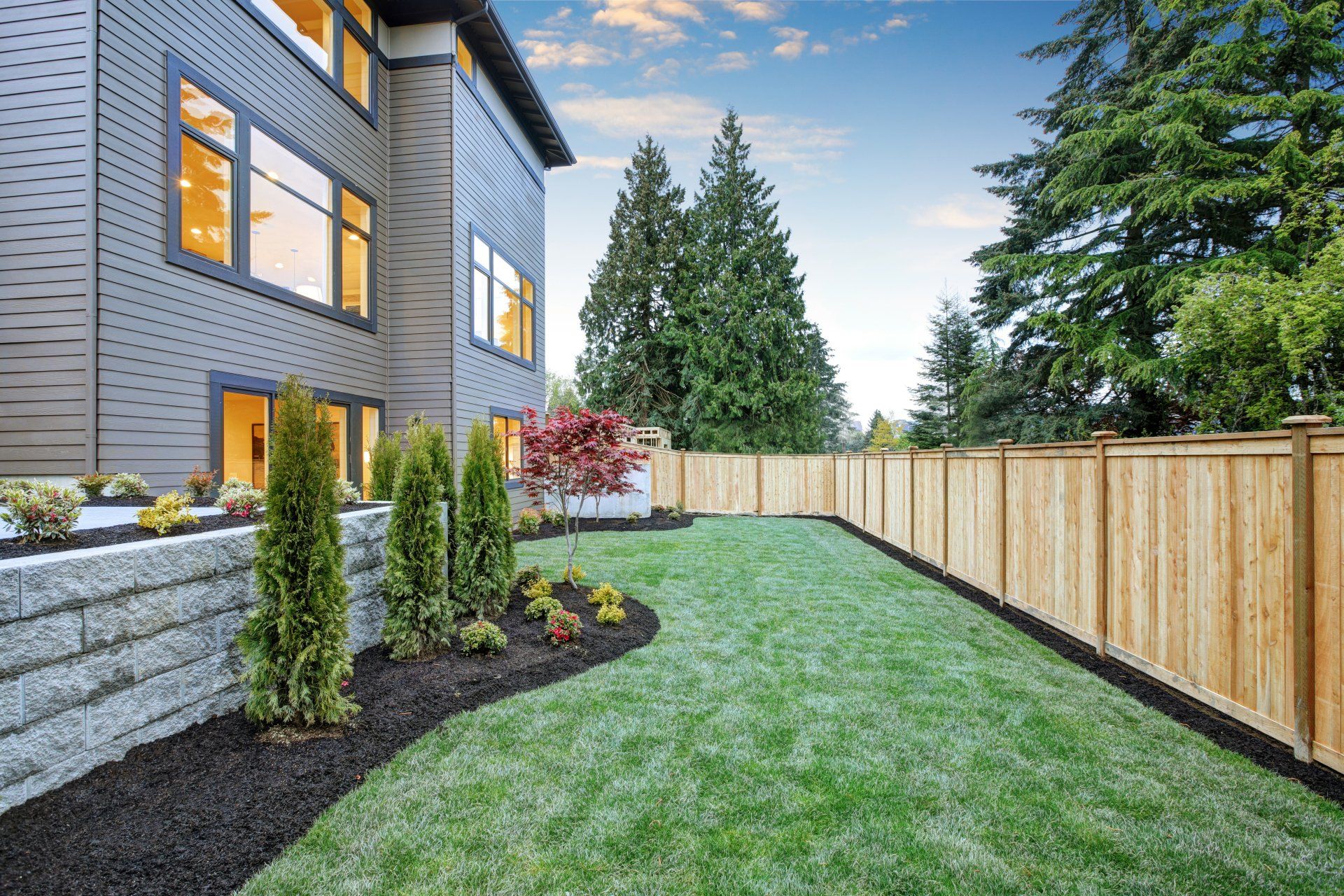 Modern home with green lawn, retaining wall, wooden fence, and evergreen trees.