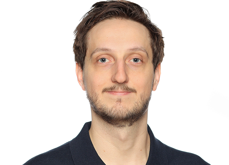 Man with light skin and short brown hair, wearing a dark blue shirt, smiling at camera against white background.