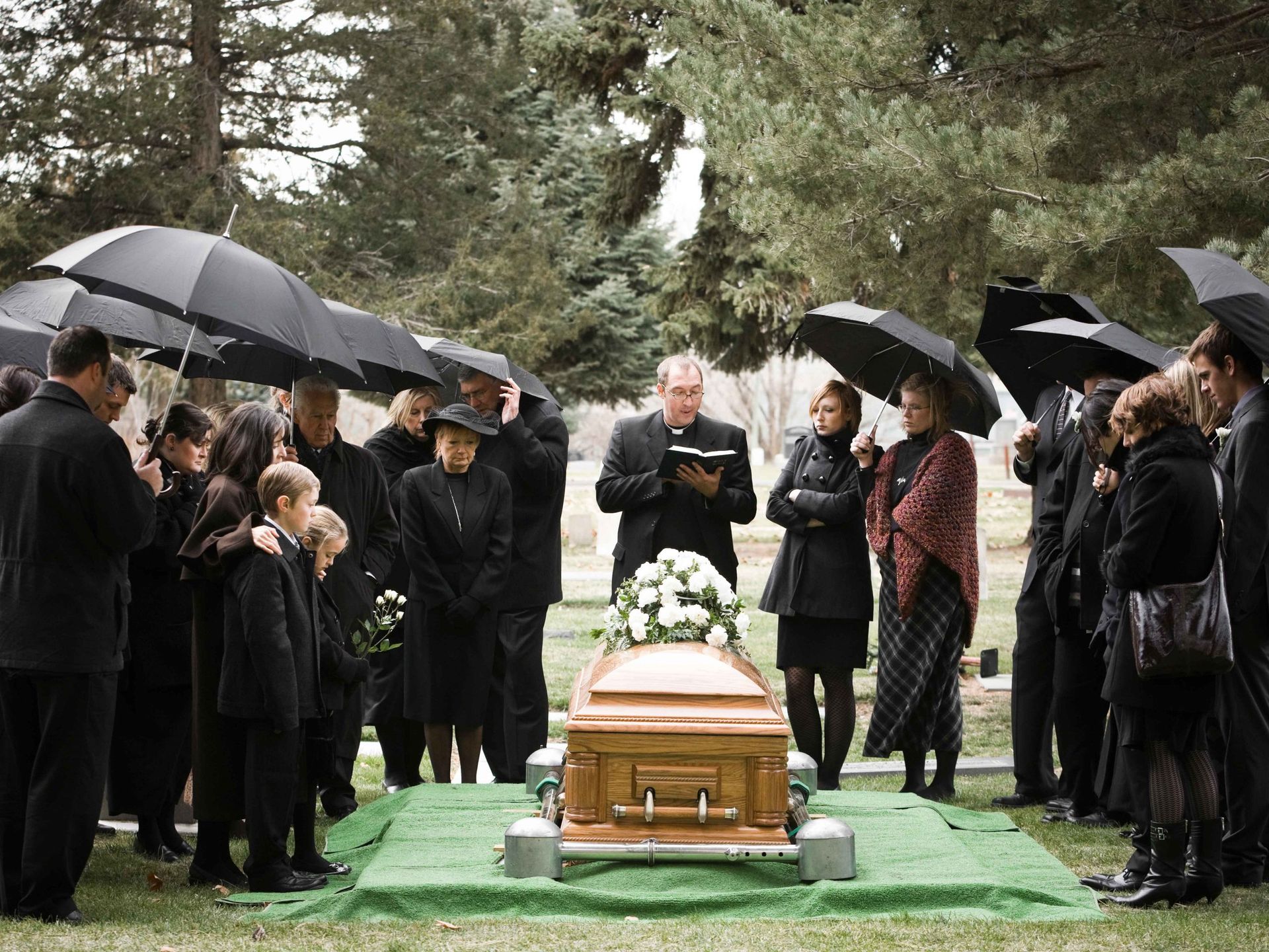 Man in black sweater at a funeral, blurred crowd in the background near flowers and a casket.