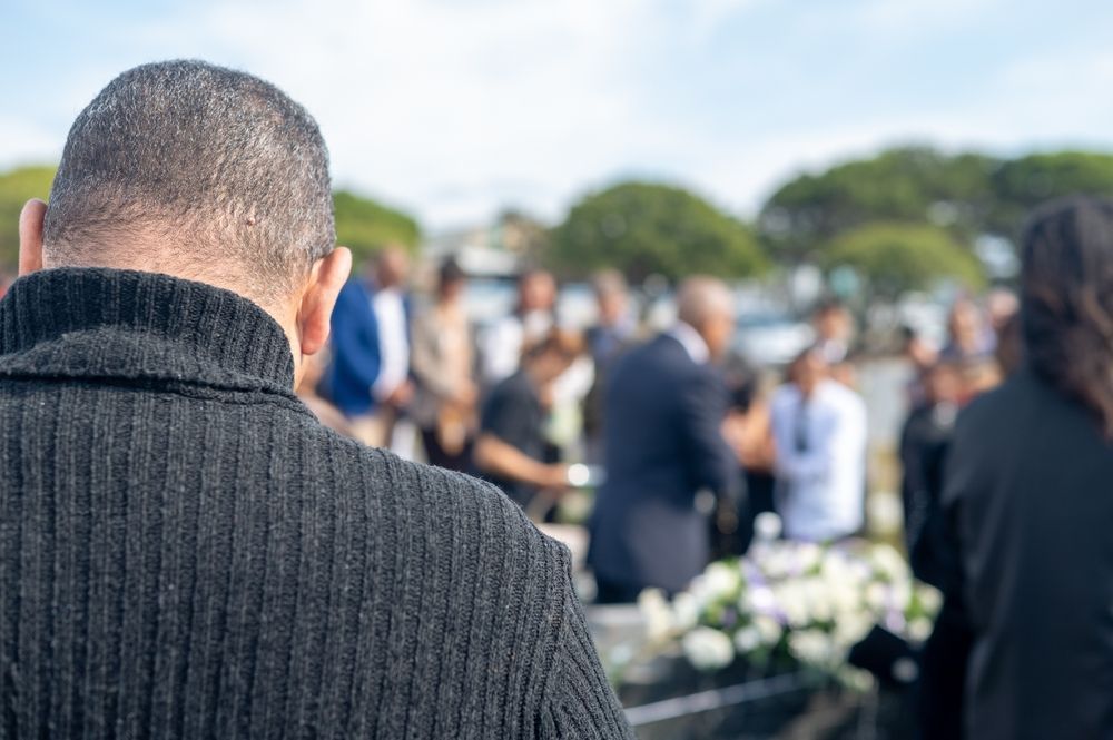 Man in black sweater at a funeral, blurred crowd in the background near flowers and a casket.