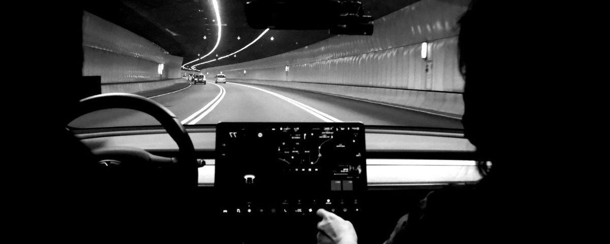 A black and white picture of a person driving a Tesla Model 3 through a tunnel.