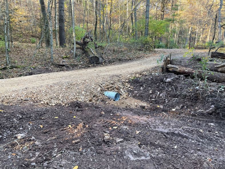 Gravel path through a wooded area with a culvert. Dirt and logs are visible along the path.