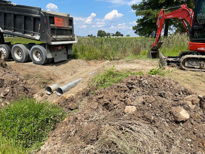 Construction site: Dump truck and excavator near field; culvert pipes in trench.