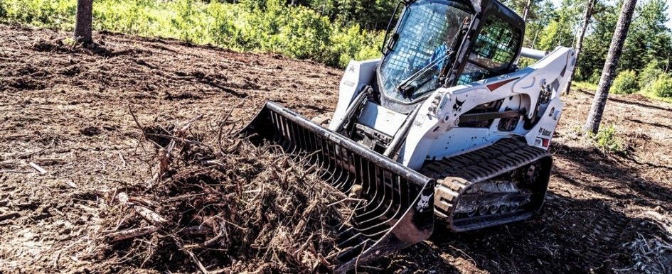 A white Bobcat skid steer with a rake attachment, clearing debris from a muddy area.