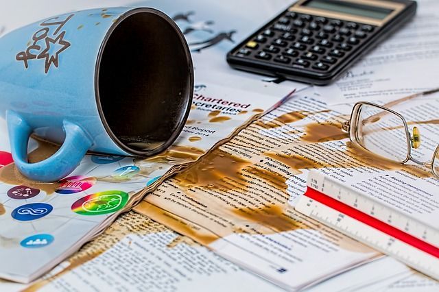 A cup of coffee spilled on a book next to a calculator and glasses.