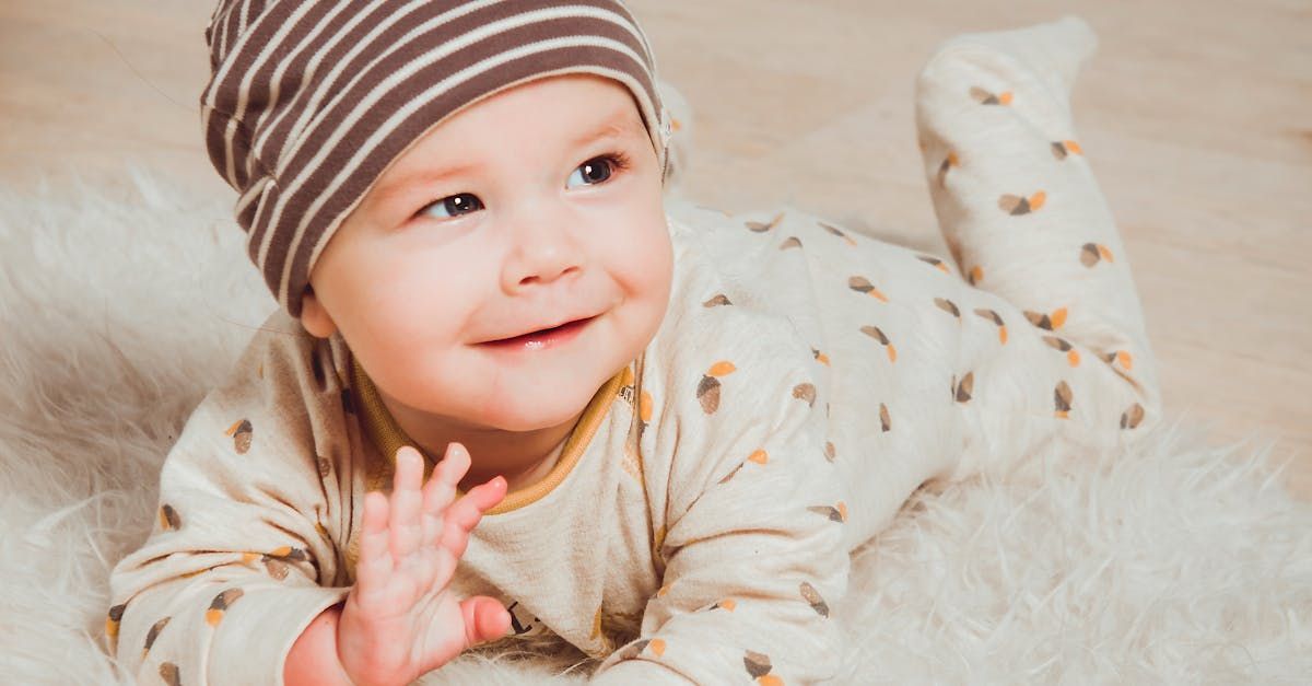 A baby wearing a striped hat is laying on a white blanket.