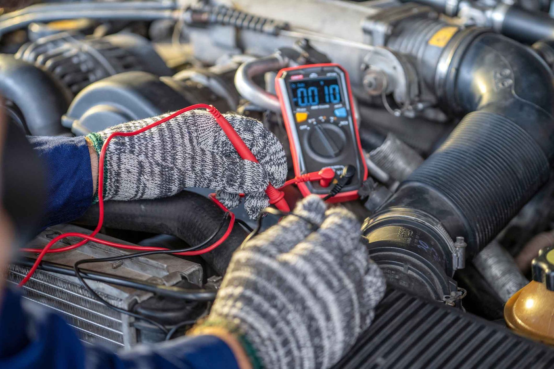 A person wearing gloves is using a multimeter to test a car engine in a well-lit setting.