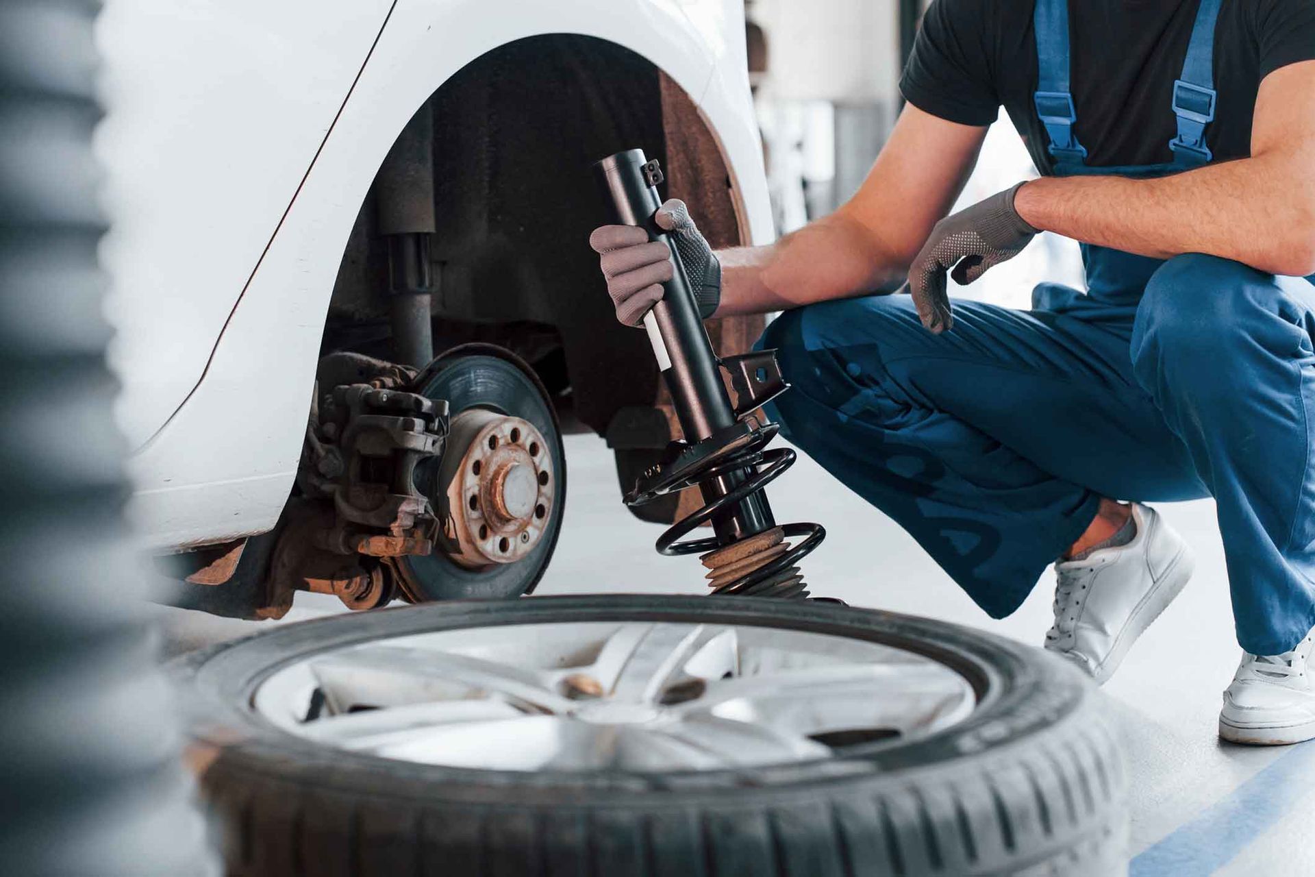 Mechanic in blue overalls replacing a shock absorber on a white car, with a spare tire in the foreground. Mechanic in blue overalls replacing a shock absorber on a white car, with a spare tire in the foreground.