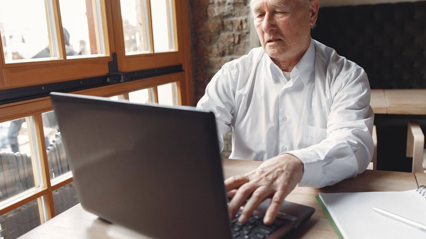 An elderly man is sitting at a table using a laptop computer.