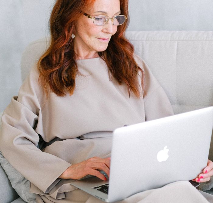 A woman wearing glasses is sitting on a couch using an apple laptop